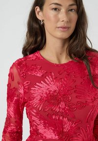 Rød kjole med blomsterbroderi, i tekstureret stof, med rund halsudskæring og lange, gennemsigtige ærmer. Designet indeholder hævede blomsterblade og roser.