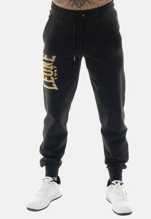 Black jogger pants with gold "LEONE 1947" text on left thigh, worn with white sneakers, waist and lower torso visible, tattooed skin above waistband.