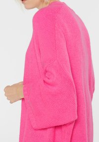 Person in einem leuchtend rosa gestrickten Pullover mit fallenden Schultern, von der Seite und bis zur Mitte des Oberkörpers vor einem schlichten Hintergrund gezeigt.
