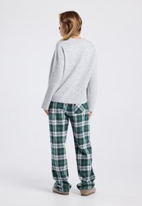 Vrouw die naar links staat, gekleed in een lichtgrijze trui, groen-witte geblokte broek en beige slippers, tegen een effen witte achtergrond.