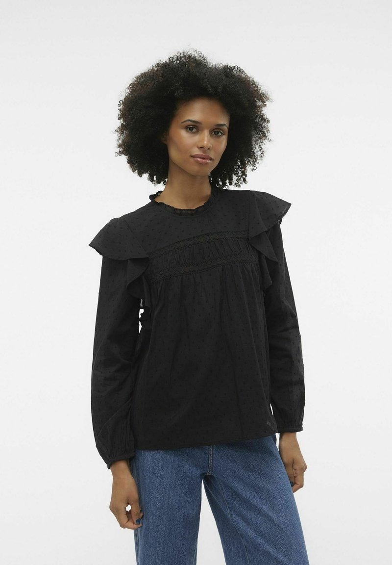 Blusa nera con spalle arricciate, tessuto testurizzato, motivo a pois irregolari e cucitura frontale arricciata; abbinata a jeans in denim blu.