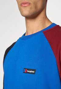 Blue and burgundy raglan sleeve t-shirt made of cotton, featuring a prominent black logo patch with red and blue accents on the chest.