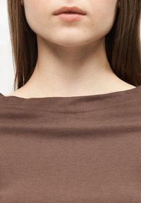 Dolna część twarzy i szyja osoby o prostych brązowych włosach, ubrana w brązową bluzkę z szerokim dekoltem na jednolitym tle.