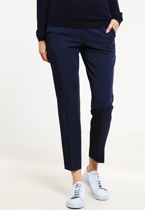 Persona che indossa pantaloni slim fit blu navy con mano in tasca e sneakers bianche, in piedi contro uno sfondo bianco uniforme.