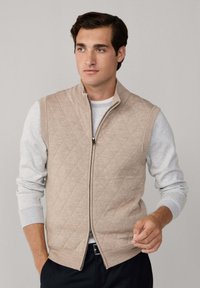 Bež preklopni prsluk s visokim ovratnikom i prednjim patentnim zatvaračem, preko svijetlo sive košulje dugih rukava. Mekana tekstura, elegantan dizajn.