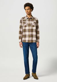 Camisa de franela a cuadros en beige, amarillo y marrón, con dos bolsillos en el pecho, combinada con jeans azules y botas marrones. Tejido texturizado con un corte clásico.