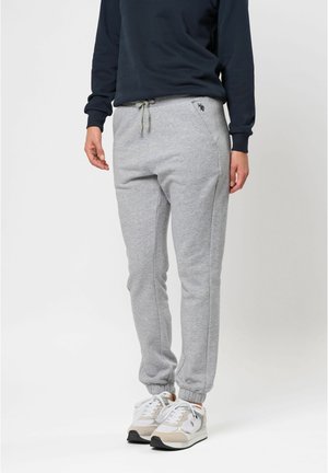 Personne portant un pantalon de survêtement gris clair avec des poignets élastiques, un haut noir à manches longues et des baskets blanches, debout sur un fond uni.