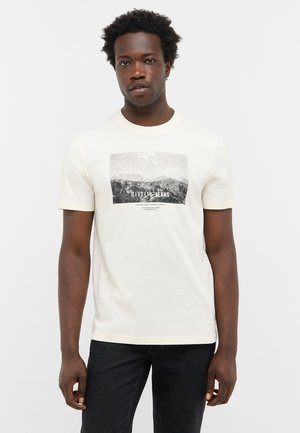 Cream short-sleeve t-shirt featuring a black and white mountain graphic with text. Soft texture, classic fit, paired with dark denim jeans.