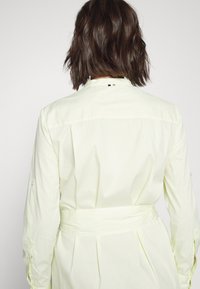 Abito camicia giallo chiaro con strisce bianche verticali, maniche lunghe, vita con cintura e colletto a punta. La parte posteriore presenta un logo del marchio discreto.