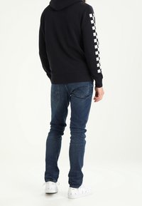 Sweat à capuche noir avec capuche et motif damier blanc le long de la manche droite, associé à un jean bleu et des baskets blanches. Coupe décontractée.