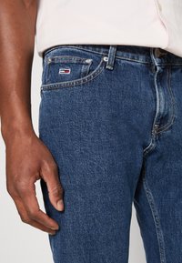 Mørkeblå denimjeans med synlige sømmer, klassisk fem-lomme-design og en liten merket etikett på frontlommen.
