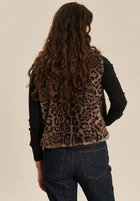 Vrouw met lang krullend haar die een leeuwenprint bontvest over een zwart shirt met lange mouwen en donkerblauwe jeans draagt, met haar rug naar de camera gekeerd.