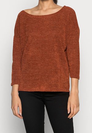 Femme portant un pull en maille texturée couleur rouille avec des manches trois-quarts et un pantalon noir, debout devant un fond uni.