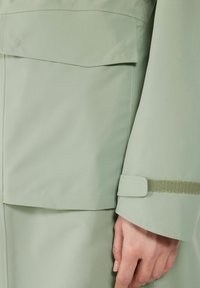 Detalle de una chaqueta impermeable verde claro que muestra un tejido suave, un bolsillo con solapa y un acento de tejido texturizado en el puño con cierre de Velcro.