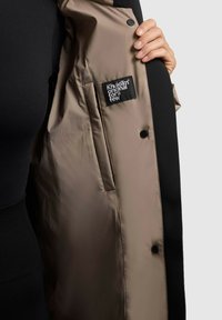 Black coat with a soft, tan lining. It features a tag that reads "Khujo original for a few" and a snap button detail on the interior.