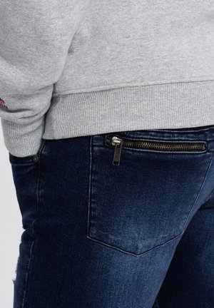 Felpa grigia con polsini a costine, abbinata a jeans in denim blu scuro con tasca laterale con cerniera e una leggera sbiaditura.