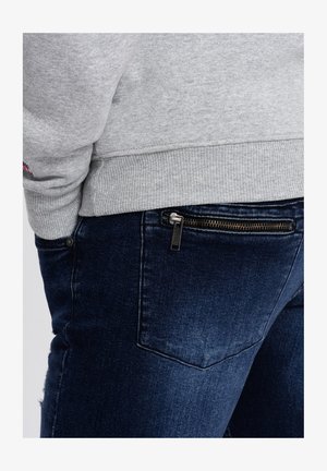 Szara bluza z ściągaczami na mankietach, zestawiona z ciemnoniebieskimi dżinsami z zasuwaną kieszenią po boku i minimalnymi przetarciami.