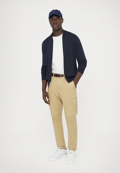 Cardigã azul-marinho canelado, t-shirt branca, calças cargo bege com bolsos e ténis brancos. Boné azul com detalhe de logótipo vermelho.