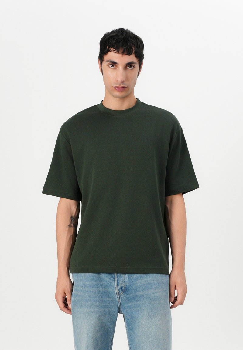 Dark green oversized t-shirt made from soft cotton, featuring a crew neckline and short sleeves. Worn with light blue denim jeans.