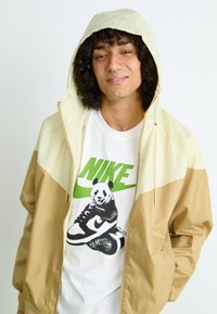 Lys beige og creme hættejakke med lynlås, båret over en hvid t-shirt med et panda- og Nike-logo i grønt.