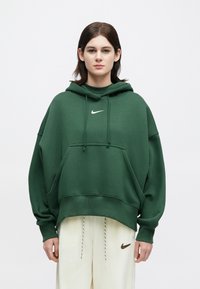 Ungdom som har på seg en mørkegrønn, oversized Nike hettegenser og kremfargede Nike joggebukser, står mot en hvit bakgrunn.