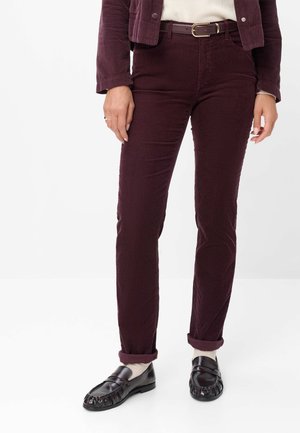 Femme portant un pantalon en velours côtelé violet foncé et une veste assortie, un haut beige, des chaussettes blanches et des mocassins noirs, debout devant un fond blanc.