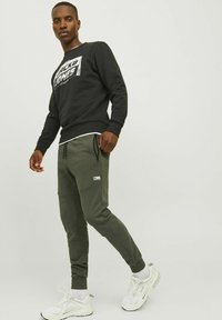 Zwarte sweatshirt met witte logo, olijfgroene taps toelopende joggers met zijzakken, en witte sportschoenen met lichte accenten.