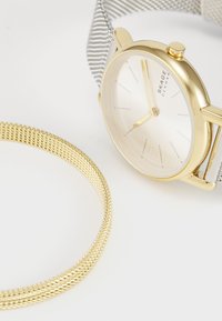 Relógio dourado com uma pulseira de malha prateada e um mostrador minimalista branco, acompanhado por uma pulseira de malha dourada combinante.