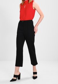 Žena v bezrukávovém červeném topu, černých širokých kalhotách s vysokým pasem a černých mules na blokovém podpatku, stojící na jednolitém pozadí.