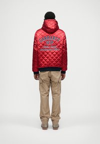 Κόκκινο καπιτονέ μπουφάν με κουκούλα, που διαθέτει κεντημένο κείμενο "CARHARTT WIP Active Jacket Anniversary Edition," σε συνδυασμό με μπεζ cargo παντελόνια.