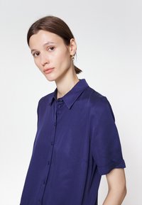 Camisa azul-marinho de manga curta com botões e colarinho, apresentando uma textura suave e dois bolsos frontais.