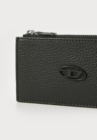 Juodas tekstūruotas odinis piniginės su užtrauktuku ir apvaliu reljefiniu logotipu priekyje. Lygūs kraštai, kompaktiška stačiakampio forma.