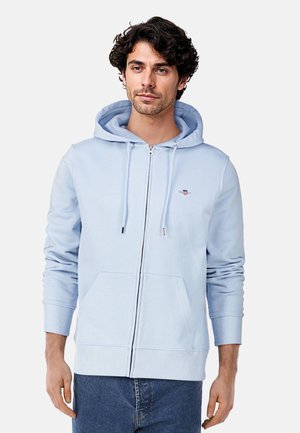 Helle blaue Zip-Hoodie aus weichem Stoff, mit einer verstellbaren Kapuze, Fronttaschen und gerippten Bündchen mit silbernen Hardware-Akzenten.