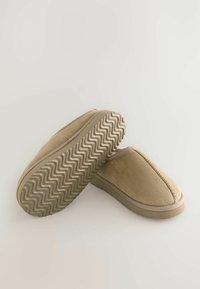 Zapatillas de ante beige con una parte superior suave y suela de goma que presenta un patrón de zigzag para mayor tracción. Punta redondeada y con un detalle de costura mínimo.