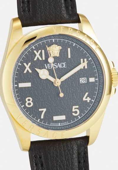 Arany színű óra fekete bőrszíjjal. Texturált szürke számlap, arany órijelzőkkel, Versace logóval és dátumablakkal a 3 órás pozícióban.