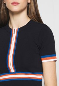 Haut côtelé noir avec une petite fermeture éclair, souligné par des rayures bleues, orange et blanches le long du col et des manches.