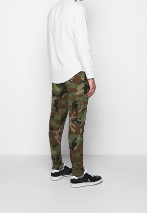 Persona vistiendo pantalones de camuflaje verde y marrón, camiseta de manga larga blanca y zapatillas negras y blancas, de pie frente a un fondo blanco liso.