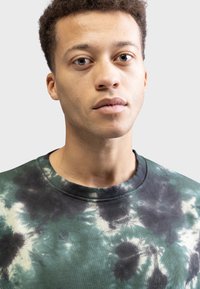 Ung mand med kort krøllet hår og alvorligt udtryk iført en grøn og sort tie-dye sweatshirt foran en ensfarvet lys baggrund.