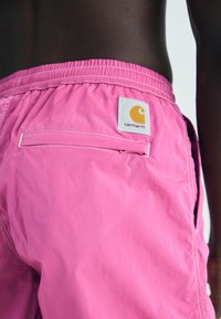 Rosa shorts laget av lett stoff med elastisk midjebånd, baklomme og et Carhartt-logo-merke i oransje på hvit bakgrunn.