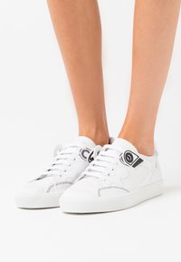 Białe skórzane trampki o teksturowanym wykończeniu, z czarnymi akcentami i wyraźnym logo obok sznurówek. Klasyczny design na sznurowadła.