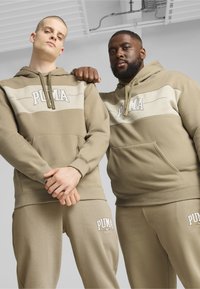 Felpe beige con cappuccio e pantaloni abbinati presentano un logo PUMA bianco. Tessuto morbido con una vestibilità comoda. Due modelli posano fianco a fianco.