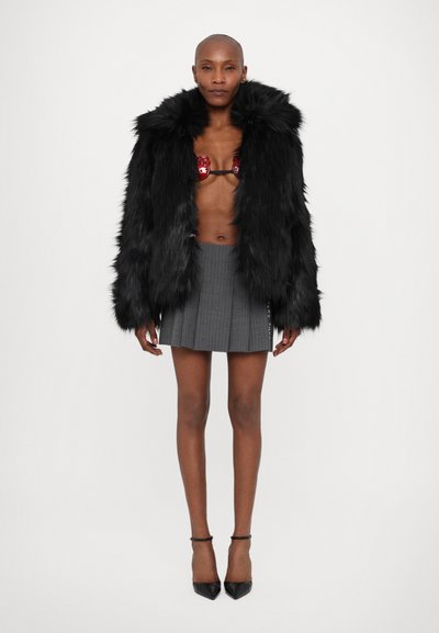 Jachetă din faux fur neagră cu guler lat, braletă detaliată cu roșu, fustă mini cu carouri gri și pantofi cu toc ascuțit negri. Texturi netede pe tot parcursul.