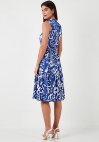 Blue and white patterned sleeveless dress with a fitted bodice and a flared, tiered skirt. Back view shows a slight scoop neckline. White heels.