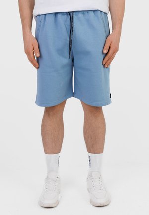 Mężczyzna w jasnoniebieskich spodenkach na sznurku, białych skarpetkach z napisem oraz białych butach sportowych, stojący na jednolitym tle.