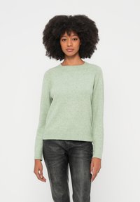 Vaaleanvihreä neulottu villapaita pyöreällä pääntiellä ja pitkillä hihoilla, valmistettu pehmeästä, teksturoidusta kankaasta. Yhdistetty mustiin farkkuihin.