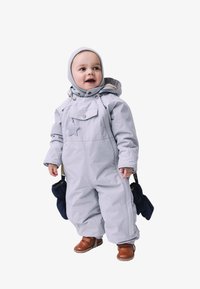 Svetlo šedý jednodielny snowsuit s zipsom, odnímateľnou kapucňou a detailom hviezdy. Nosí sa s hnedými bootami a tmavomodrými rukavicami, stojaci postoj.