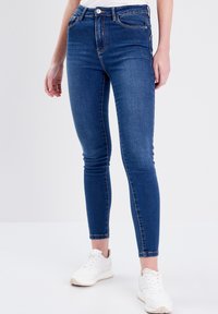 Jeans ajustados en denim azul oscuro, con cintura alta, piernas rectas, sutiles desgastes y diseño clásico de cinco bolsillos. Combinados con zapatillas blancas.