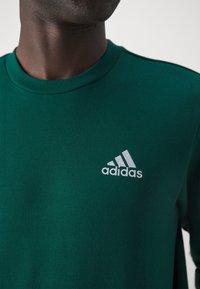Zelená bavlněná mikina s kulatým výstřihem, která na levé straně hrudi nese šedé logo Adidas, má hladkou texturu a klasický střih.