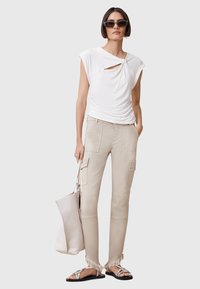 Top blanco ajustado con un detalle de nudo, pantalones cargo beige con bolsillos y sandalias plateadas con tiras. La modelo sostiene un gran bolso tote beige.