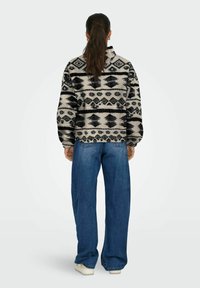 Pull en polaire noir et crème à motifs avec un col montant, associé à un jean bleu à jambes larges et des baskets blanches.